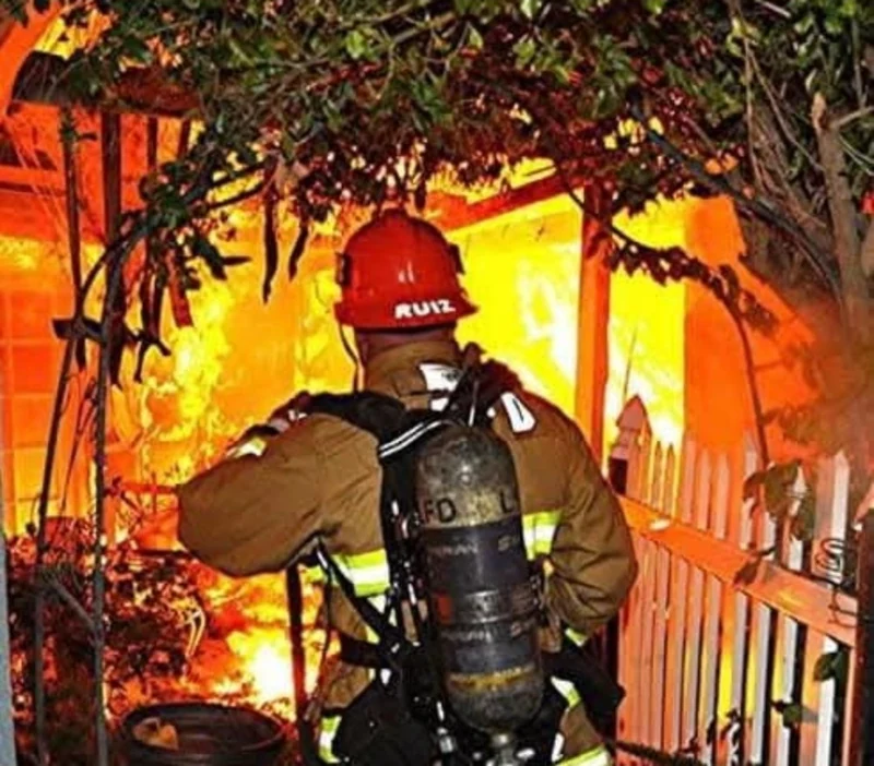 A firefighter wearing a red helmet and breathing apparatus in front of a burning structure.